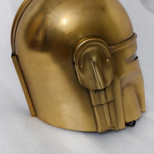 May include: A gold-colored helmet with a rounded top and a visor area. The helmet has a smooth, metallic finish and features detailed side panels and a chin piece. The overall design suggests a futuristic or sci-fi theme.