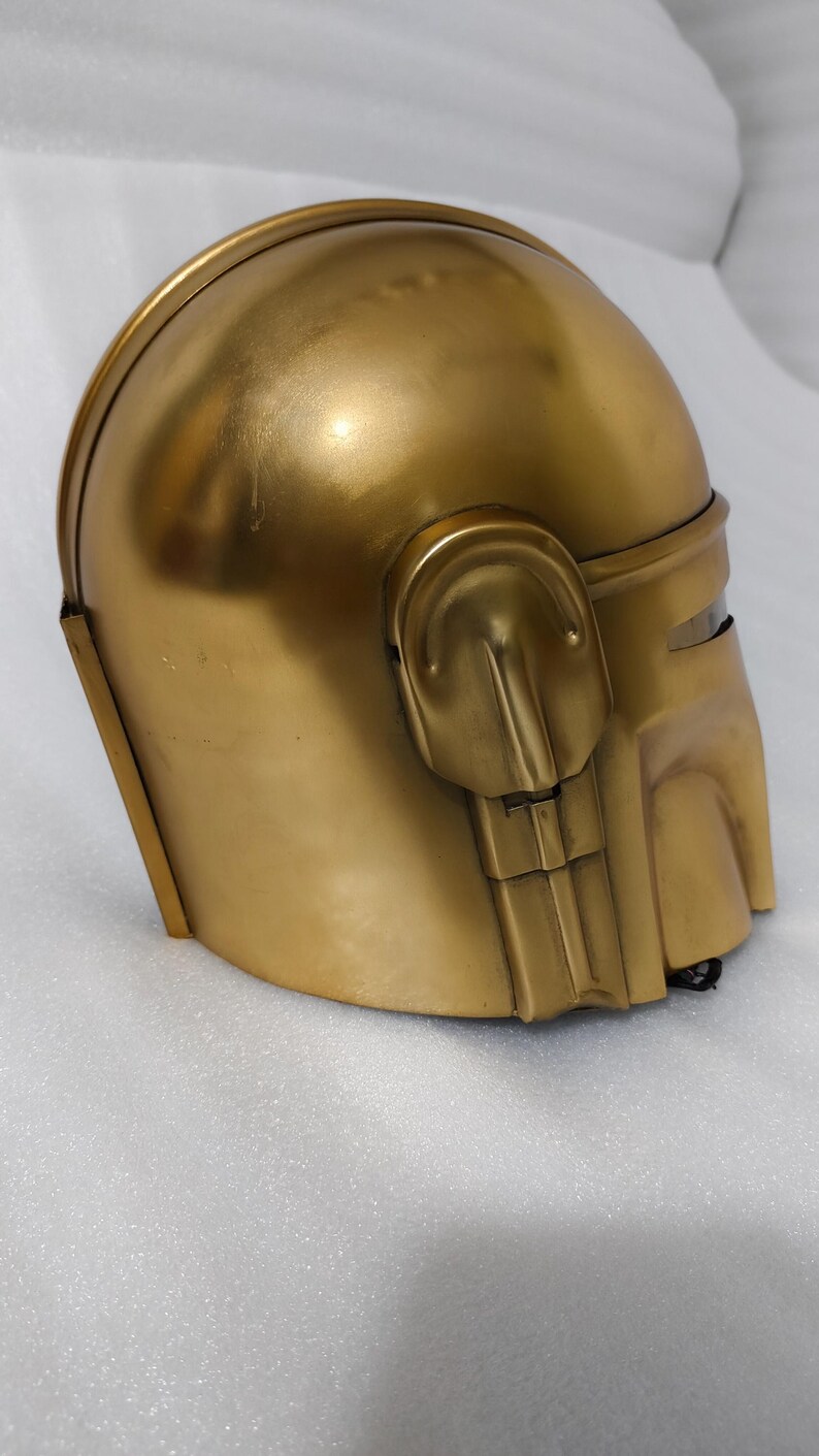 May include: A gold-colored helmet with a rounded top and a visor area. The helmet has a smooth, metallic finish and features detailed side panels and a chin piece. The overall design suggests a futuristic or sci-fi theme.