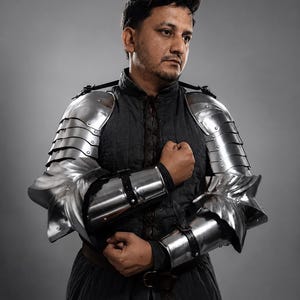Puede incluir: Una persona con armadura de estilo medieval, que incluye hombreras y piezas de brazo articuladas de acero. La armadura plateada contrasta con la tela oscura de la prenda debajo. La persona posa sobre un fondo gris.