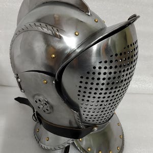 May include: A polished steel helmet with a visor featuring numerous small holes. The helmet has a rounded crown, riveted details, and a leather strap. The design suggests medieval or historical armor.