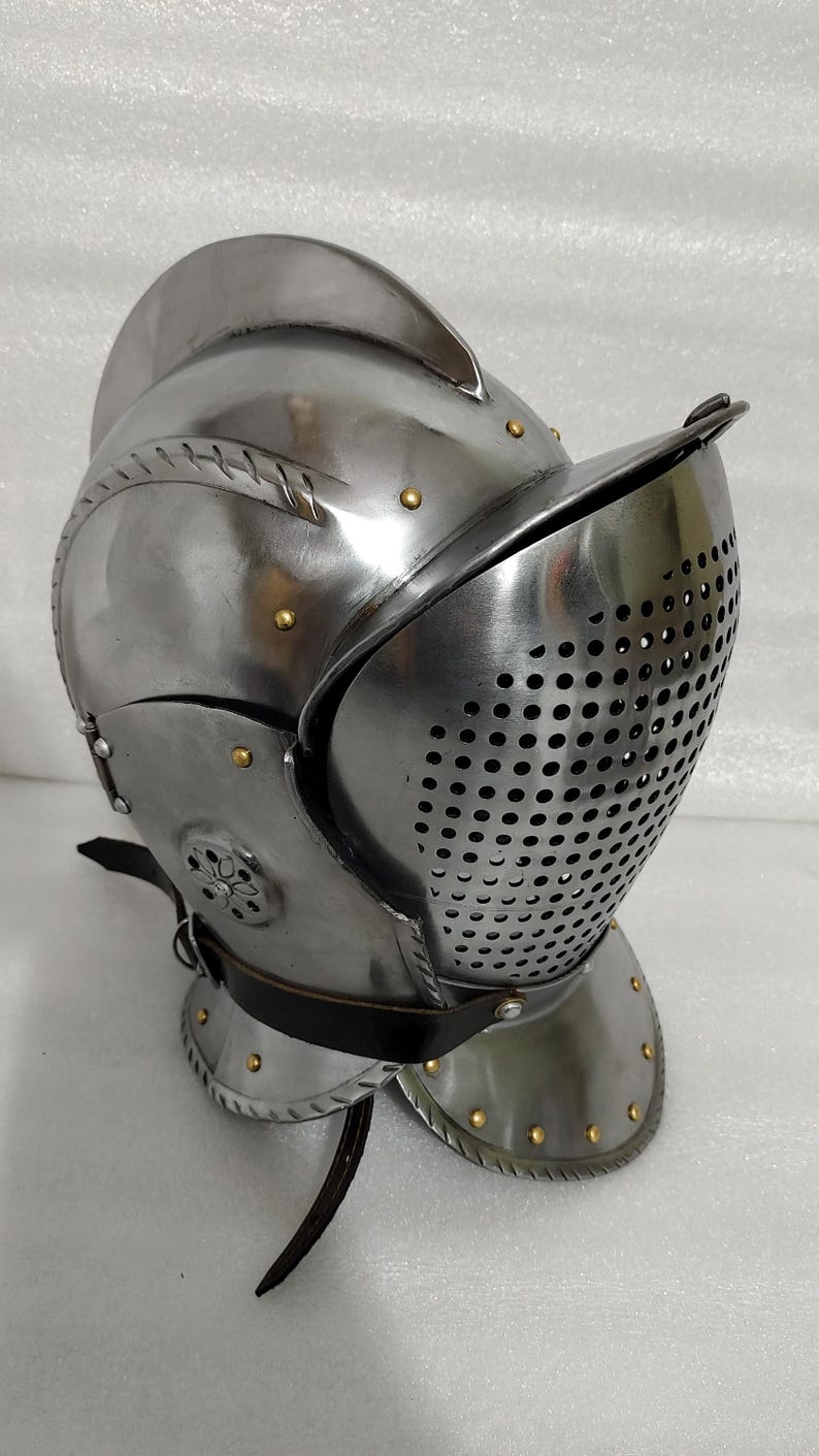 May include: A polished steel helmet with a visor featuring numerous small holes. The helmet has a rounded crown, riveted details, and a leather strap. The design suggests medieval or historical armor.