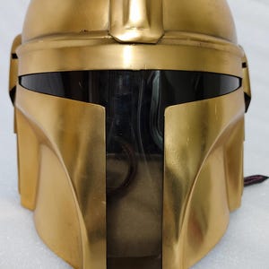 May include: A gold-colored helmet with a rounded top and a visor. The helmet has a smooth, metallic finish and a distinctive shape, with a raised ridge running along the top. The visor is black, and the helmet appears to be a prop or costume piece.