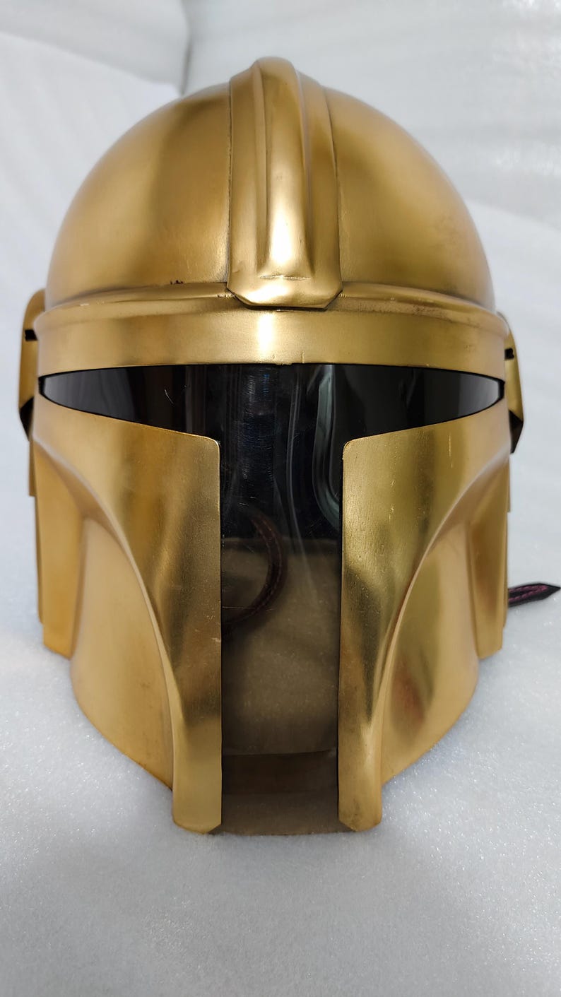 May include: A gold-colored helmet with a rounded top and a visor. The helmet has a smooth, metallic finish and a distinctive shape, with a raised ridge running along the top. The visor is black, and the helmet appears to be a prop or costume piece.