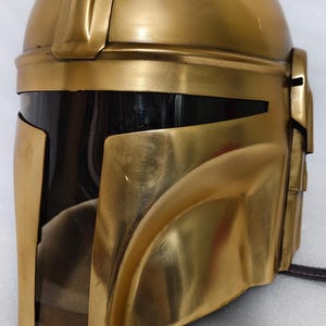 May include: A gold-colored helmet with a dark visor, likely a costume piece. The helmet has a smooth, metallic finish and a distinctive shape, with a rounded top and angled cheek pieces. The visor is black, and the overall design suggests a futuristic or sci-fi theme.