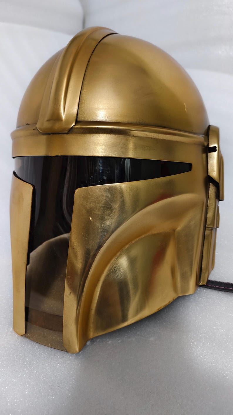 May include: A gold-colored helmet with a dark visor, likely a costume piece. The helmet has a smooth, metallic finish and a distinctive shape, with a rounded top and angled cheek pieces. The visor is black, and the overall design suggests a futuristic or sci-fi theme.