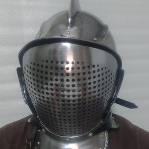 May include: A polished steel knight's helmet with a perforated visor, a prominent central ridge, and a neck guard. The helmet is secured with leather straps and rivets. The overall design suggests medieval armor.