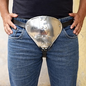 Pode incluir: Um dispositivo de castidade de metal prateado com a palavra "END" gravada. O dispositivo está preso a um cinto de couro preto e é usado sobre jeans azuis. A pessoa está vestindo uma camiseta preta.