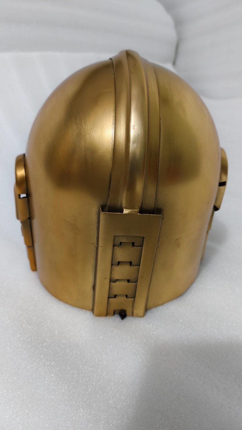 May include: A gold-colored helmet with a rounded top and vertical ridges. The helmet has rectangular and segmented side pieces and a rectangular back piece with horizontal indentations. The surface has a metallic sheen.