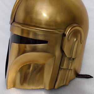 May include: A gold-colored helmet with a sleek, metallic finish. The helmet features a visor with a dark, rectangular opening and detailed side panels. The overall design suggests a futuristic or sci-fi theme.