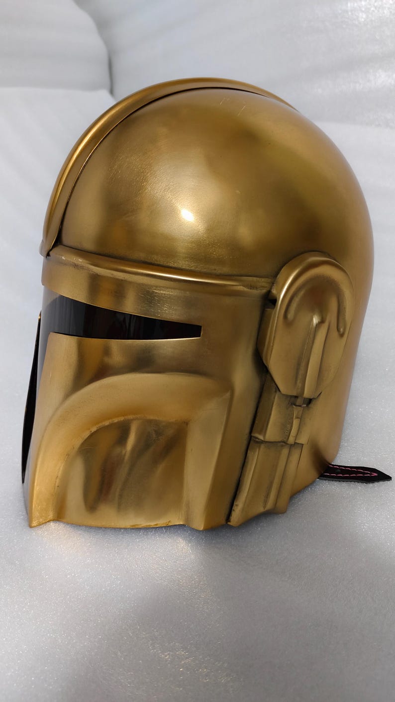 May include: A gold-colored helmet with a sleek, metallic finish. The helmet features a visor with a dark, rectangular opening and detailed side panels. The overall design suggests a futuristic or sci-fi theme.