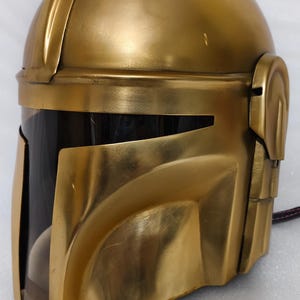 May include: A gold-colored helmet with a visor, likely a costume piece. The helmet has a smooth, metallic finish and a design with angular features. The visor is dark, and the overall shape suggests a futuristic or sci-fi theme.