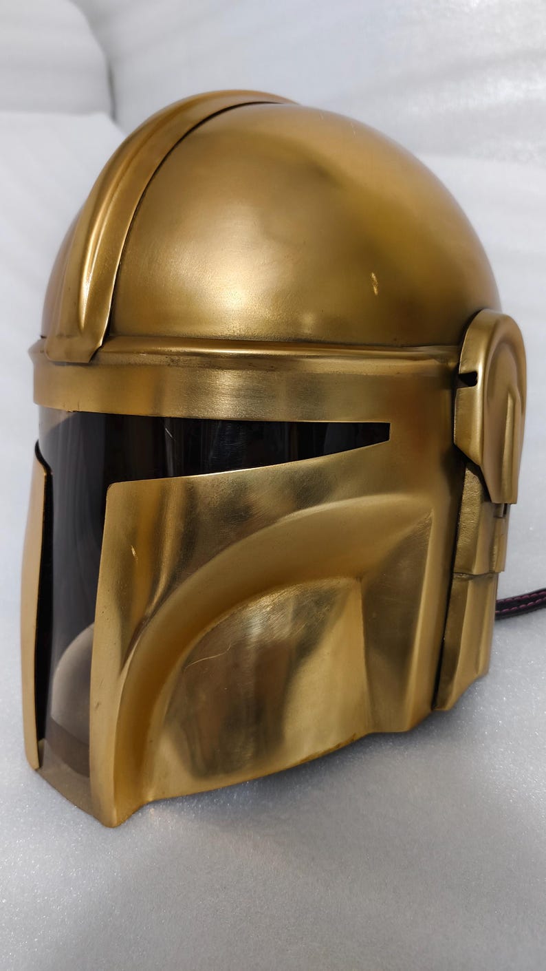 May include: A gold-colored helmet with a visor, likely a costume piece. The helmet has a smooth, metallic finish and a design with angular features. The visor is dark, and the overall shape suggests a futuristic or sci-fi theme.