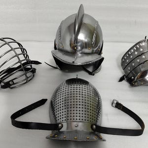 May include: Four pieces of medieval armor are displayed. The collection includes a metal helmet with a central ridge, a perforated helmet, a face guard, and a metal arm guard. All pieces are silver in color and have black leather straps.