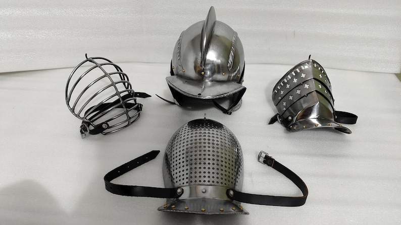 May include: Four pieces of medieval armor are displayed. The collection includes a metal helmet with a central ridge, a perforated helmet, a face guard, and a metal arm guard. All pieces are silver in color and have black leather straps.