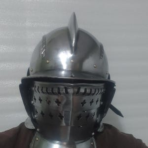 May include: A polished steel helmet with a pointed top and a visor with cross-shaped cutouts. The helmet includes a neck guard with rivets, suggesting medieval armor. The overall design is metallic and reflective.