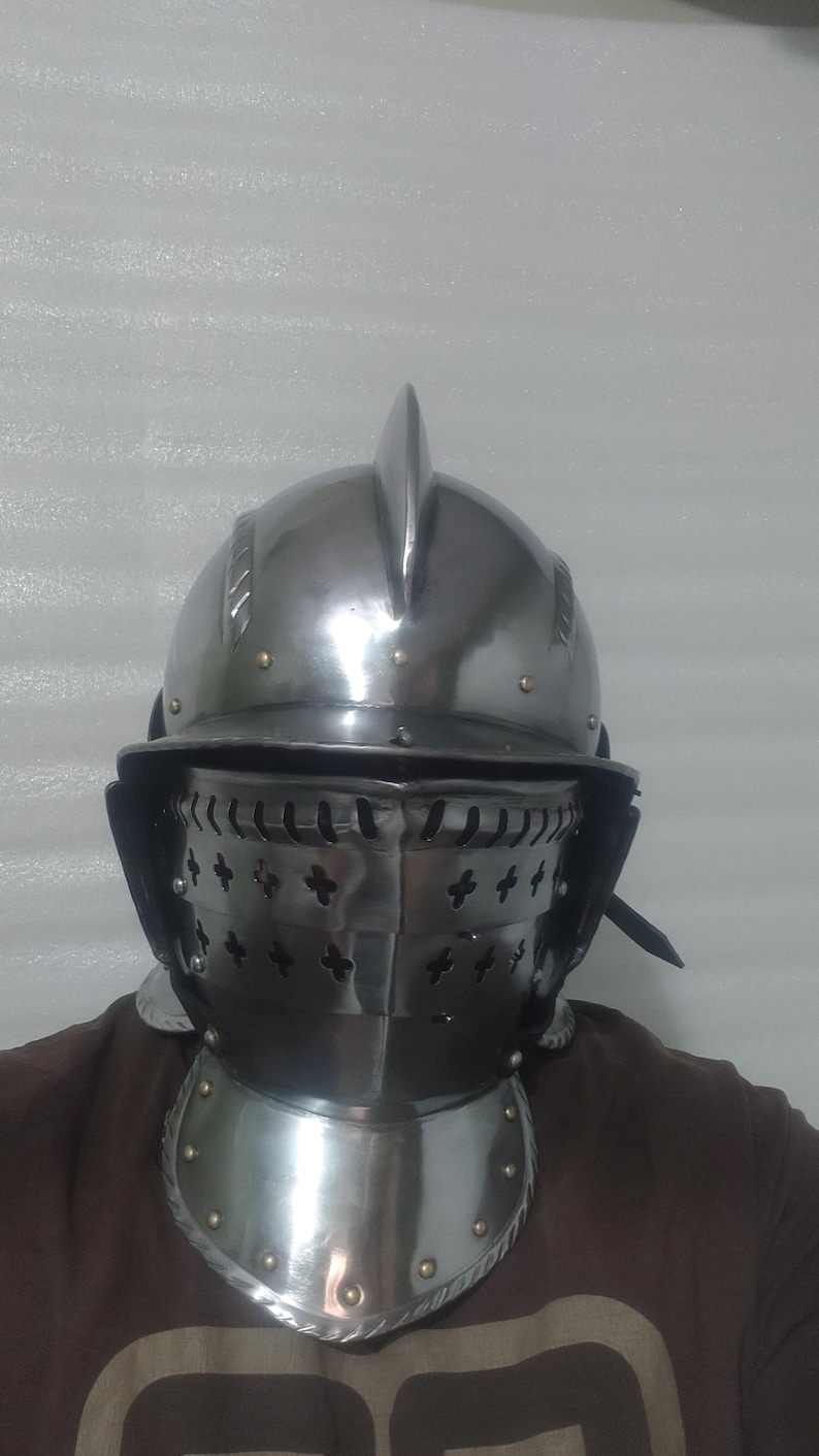 May include: A polished steel helmet with a pointed top and a visor with cross-shaped cutouts. The helmet includes a neck guard with rivets, suggesting medieval armor. The overall design is metallic and reflective.