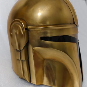 May include: A gold-colored helmet with a visor, likely for cosplay or display. The helmet has a smooth, metallic finish and a design with angular features. The visor is black, contrasting with the gold. The helmet is a replica of a popular character's helmet.