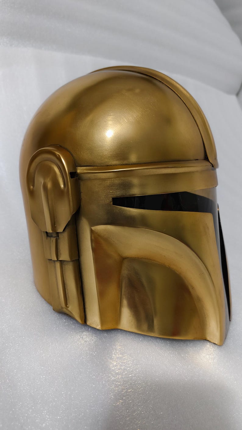 May include: A gold-colored helmet with a visor, likely for cosplay or display. The helmet has a smooth, metallic finish and a design with angular features. The visor is black, contrasting with the gold. The helmet is a replica of a popular character's helmet.