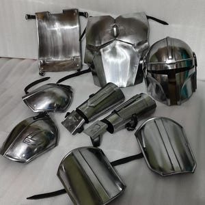 Puede incluir: Un conjunto de piezas de armadura mandaloriana plateada met&aacute;lica, que incluye un casco, una placa para el pecho, hombreras y guanteletes. La armadura tiene un acabado de metal cepillado y se muestra sobre una superficie de color claro. El conjunto est&aacute; dise&ntilde;ado para cosplay o disfraces.