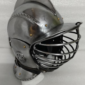 May include: A polished steel knight's helmet with a visor and a rounded top. The helmet features riveted construction and brass accents. The visor has a series of vertical bars for protection. The helmet is designed for historical reenactment or display.
