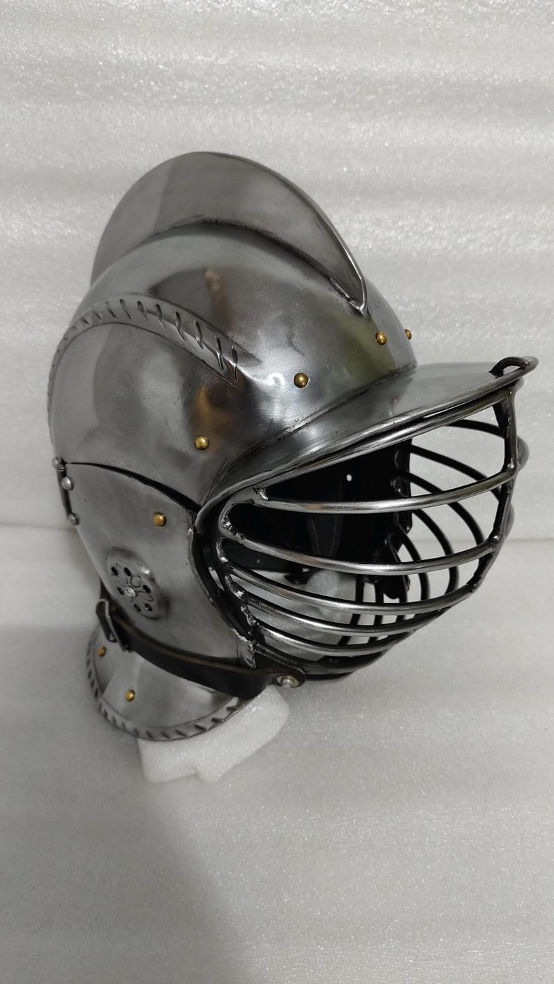 May include: A polished steel knight's helmet with a visor and a rounded top. The helmet features riveted construction and brass accents. The visor has a series of vertical bars for protection. The helmet is designed for historical reenactment or display.