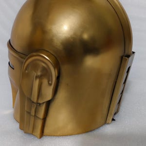 May include: A gold-colored helmet with a rounded top and angular features. The helmet has a smooth, metallic finish and includes side details and a visor area. The overall design suggests a futuristic or fantasy theme.