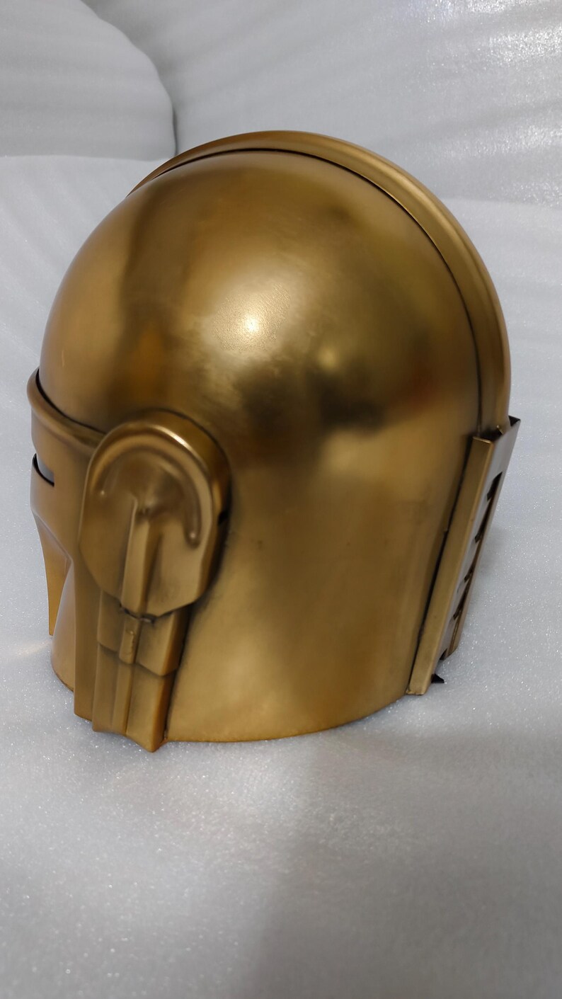 May include: A gold-colored helmet with a rounded top and angular features. The helmet has a smooth, metallic finish and includes side details and a visor area. The overall design suggests a futuristic or fantasy theme.