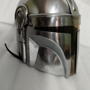 Puede incluir: Un casco plateado met&aacute;lico con una parte superior redondeada y elementos angulares. El casco tiene una visera y un dise&ntilde;o distintivo, probablemente para cosplay o exhibici&oacute;n. La superficie refleja la luz, d&aacute;ndole un aspecto pulido.