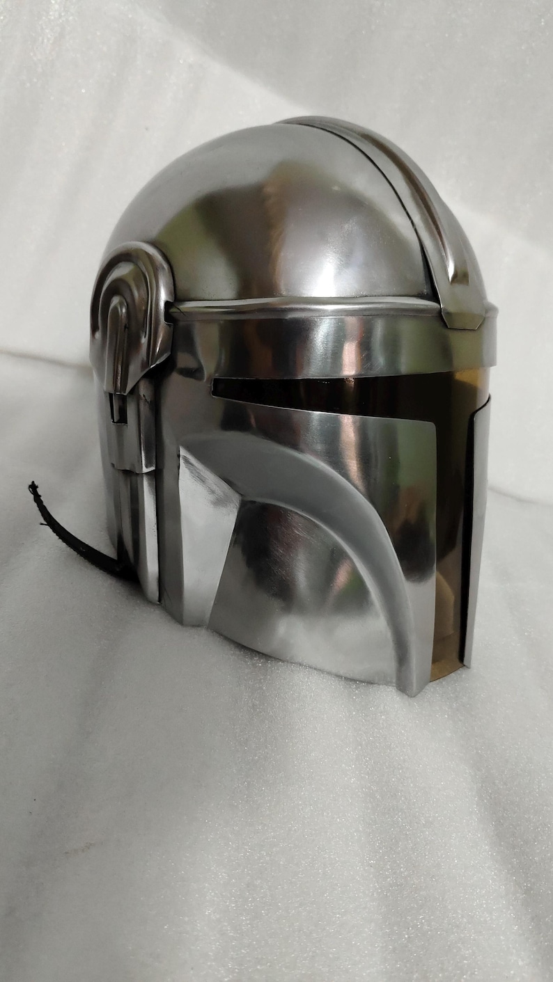 Puede incluir: Un casco plateado met&aacute;lico con una parte superior redondeada y elementos angulares. El casco tiene una visera y un dise&ntilde;o distintivo, probablemente para cosplay o exhibici&oacute;n. La superficie refleja la luz, d&aacute;ndole un aspecto pulido.