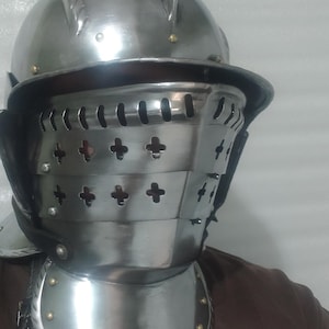 May include: A polished steel helmet with a visor and gorget. The helmet features a rounded top with a crest, a hinged visor with cross-shaped cutouts, and a neck guard. The metal armor is secured with rivets and leather straps.