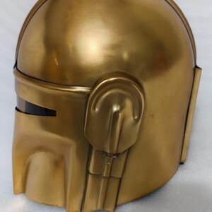 May include: A gold-colored helmet with a rounded top and angular features. The helmet has a visor and ear-like protrusions on the sides. The surface has a metallic sheen.
