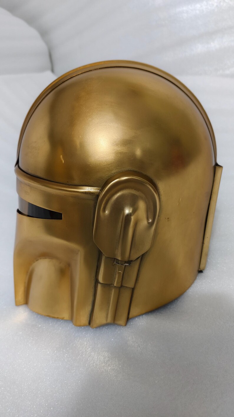 May include: A gold-colored helmet with a rounded top and angular features. The helmet has a visor and ear-like protrusions on the sides. The surface has a metallic sheen.