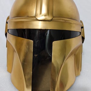 May include: A gold-colored helmet with a dark visor. The helmet has a rounded top and angular features, with a prominent ridge running down the center. The visor is dark, and the helmet appears to be made of a metallic material. The overall design suggests a futuristic or sci-fi theme.
