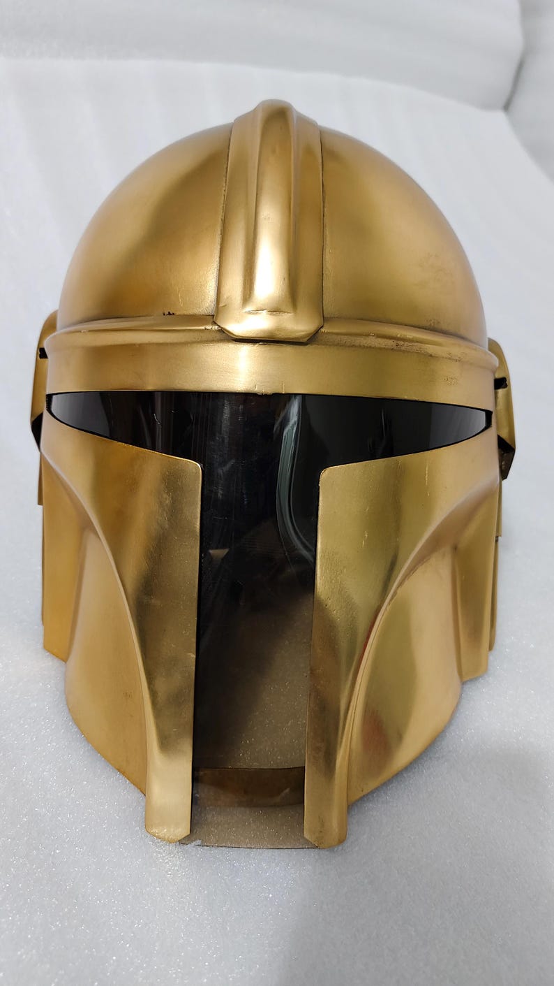 May include: A gold-colored helmet with a dark visor. The helmet has a rounded top and angular features, with a prominent ridge running down the center. The visor is dark, and the helmet appears to be made of a metallic material. The overall design suggests a futuristic or sci-fi theme.