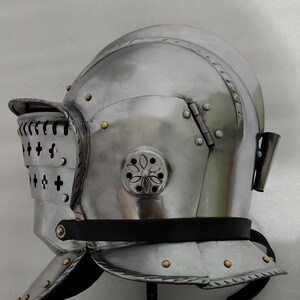 May include: A polished, silver-colored medieval helmet with a visor and neck guard. The helmet features rivets, decorative floral accents, and a black leather strap. The visor has vertical slits and cross-shaped cutouts.