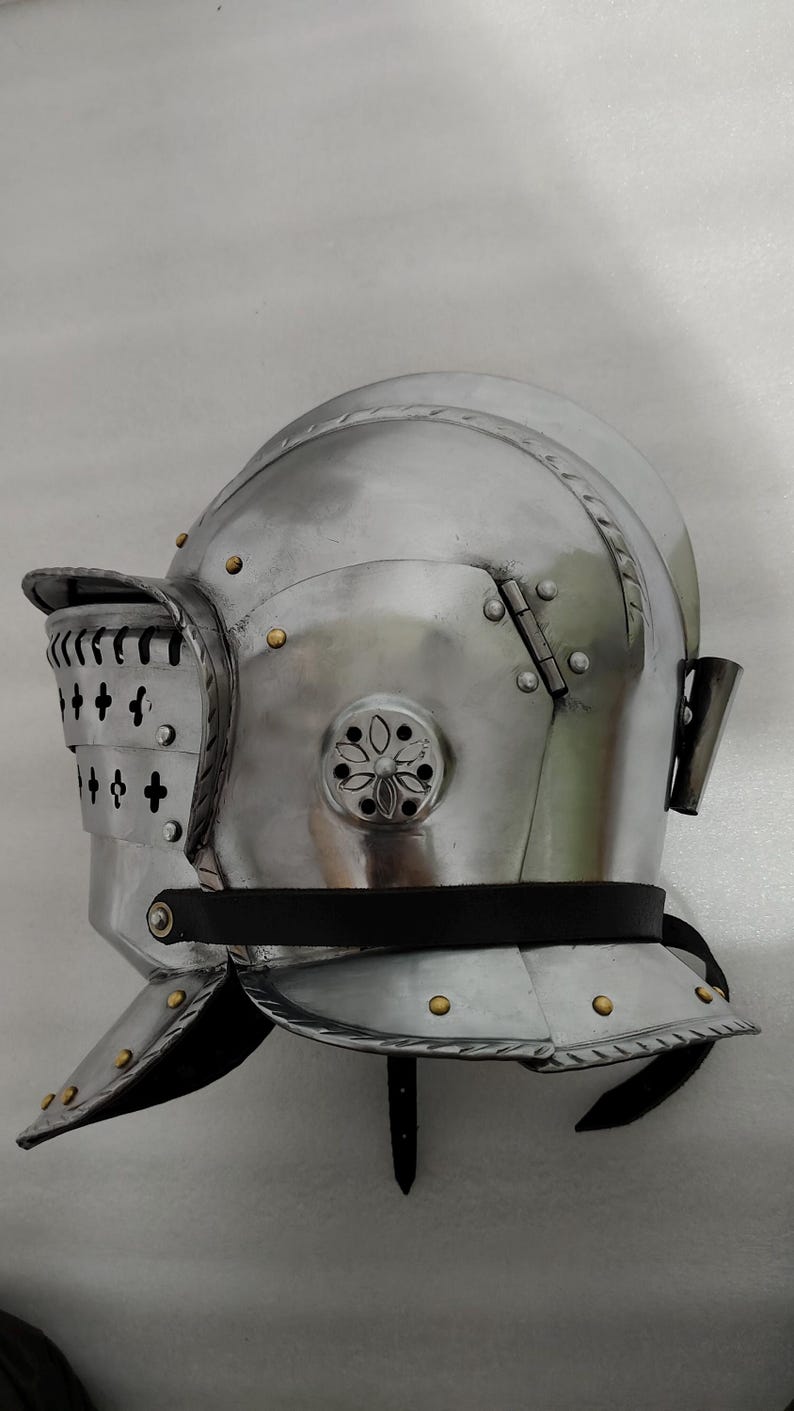 May include: A polished, silver-colored medieval helmet with a visor and neck guard. The helmet features rivets, decorative floral accents, and a black leather strap. The visor has vertical slits and cross-shaped cutouts.