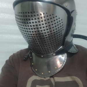 May include: A silver-colored medieval helmet with a perforated visor and a neck guard. The helmet has a rounded top with a small fin and is secured with a leather strap. The neck guard is adorned with rivets.