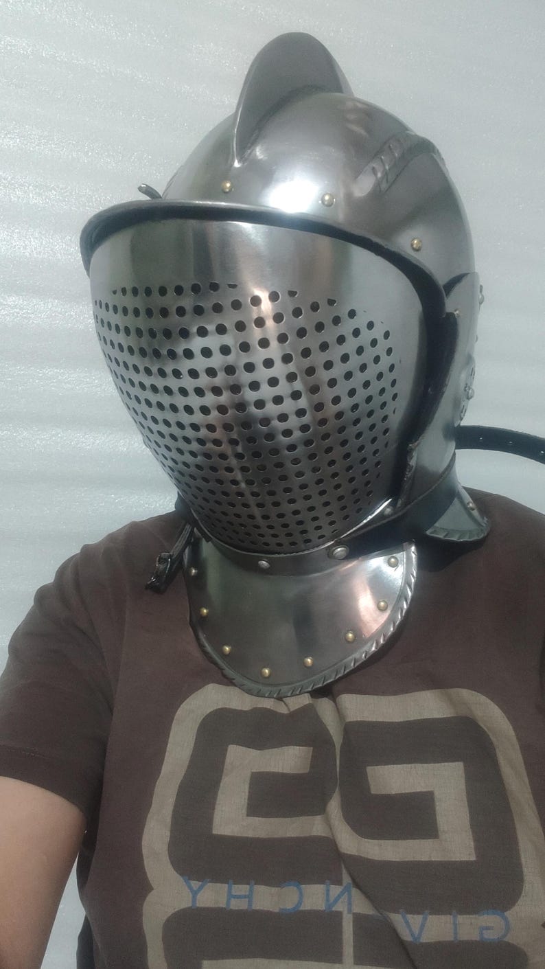 May include: A silver-colored medieval helmet with a perforated visor and a neck guard. The helmet has a rounded top with a small fin and is secured with a leather strap. The neck guard is adorned with rivets.