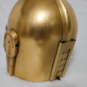 May include: A gold-colored helmet with a rounded top and a detailed side panel. The helmet has a smooth, metallic finish and a design that suggests a futuristic or retro aesthetic. The helmet's design includes a side ear piece and a series of vertical lines.