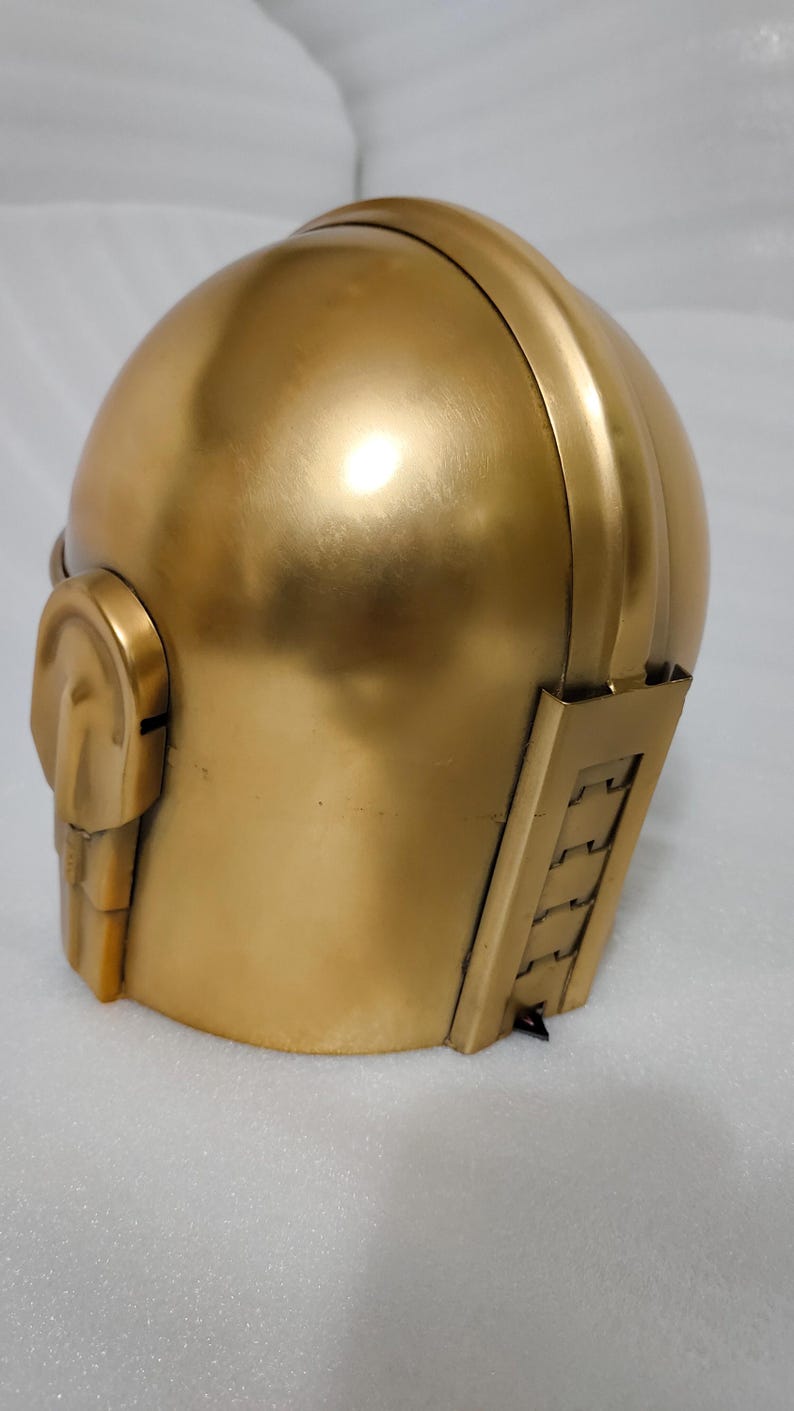 May include: A gold-colored helmet with a rounded top and a detailed side panel. The helmet has a smooth, metallic finish and a design that suggests a futuristic or retro aesthetic. The helmet's design includes a side ear piece and a series of vertical lines.