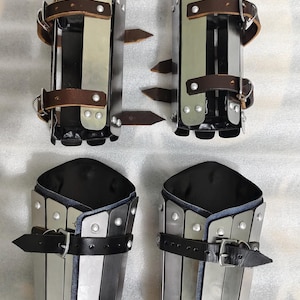 May include: Set of four pieces of medieval armor, including two greaves and two arm guards. The armor is made of metal with leather straps and rivets. The arm guards have a black interior and leather straps with buckles.