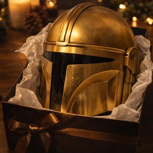 May include: A gold-colored helmet, presented in a gift box with tissue paper and a brown ribbon. The helmet has a distinctive design with a visor and is labeled "The Ultimate Gift".