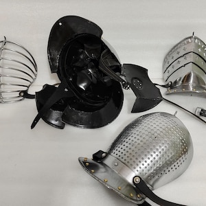 May include: An assortment of medieval helmets and face guards, including a black helmet, a silver cage-style face guard, and a silver helmet with decorative rivets. The pieces are arranged on a white surface.