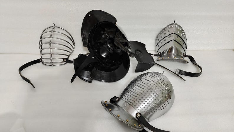May include: An assortment of medieval helmets and face guards, including a black helmet, a silver cage-style face guard, and a silver helmet with decorative rivets. The pieces are arranged on a white surface.