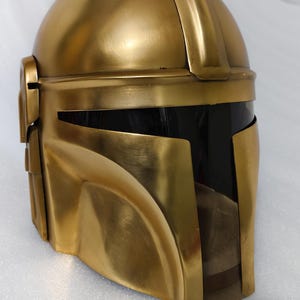 May include: A gold-colored helmet with a visor. The helmet has a rounded top and a distinctive, angular design. The visor is black, and the helmet appears to be made of metal or a similar material. The overall aesthetic suggests a futuristic or science fiction theme.