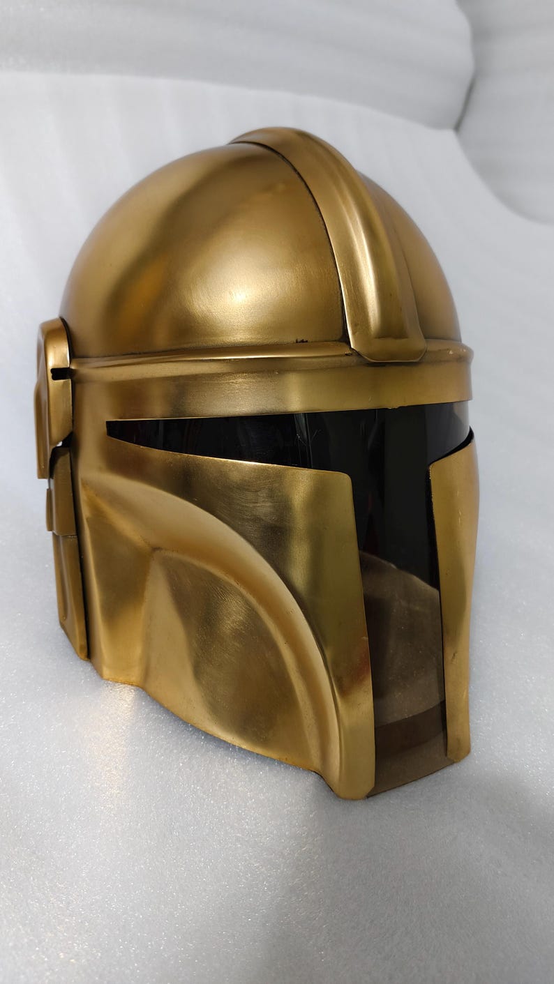 May include: A gold-colored helmet with a visor. The helmet has a rounded top and a distinctive, angular design. The visor is black, and the helmet appears to be made of metal or a similar material. The overall aesthetic suggests a futuristic or science fiction theme.