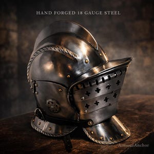 Puede incluir: Un casco de acero forjado a mano, calibre 18, con visera y remaches decorativos. El casco tiene un acabado metálico oscuro y está diseñado con detalles intrincados. El texto "HAND FORGED 18 GAUGE STEEL" es visible.