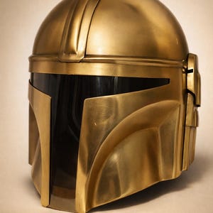 May include: A gold-colored helmet with a sleek, metallic finish. The helmet features a rounded top with a series of ridges and a visor area with a dark, reflective surface. The design includes angular cheek and jaw pieces, suggesting a futuristic or sci-fi theme.