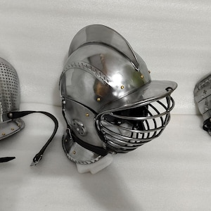 May include: Three pieces of medieval armor, including a helmet with a visor, a perforated helmet, and a shoulder guard. All pieces are made of shiny, silver-colored metal and feature black leather straps.