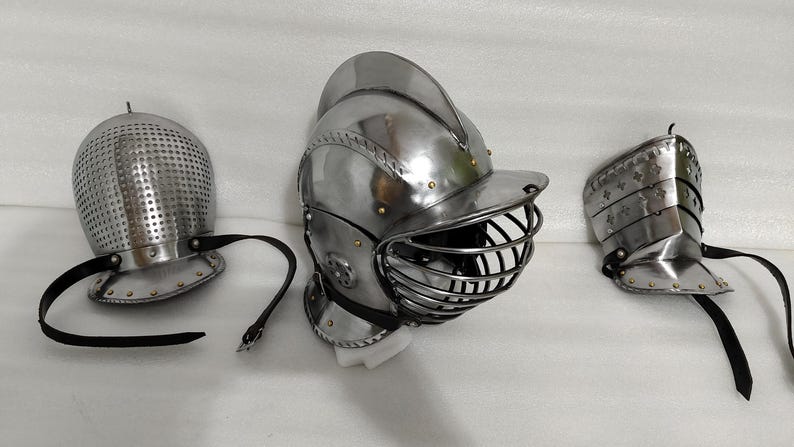 May include: Three pieces of medieval armor, including a helmet with a visor, a perforated helmet, and a shoulder guard. All pieces are made of shiny, silver-colored metal and feature black leather straps.